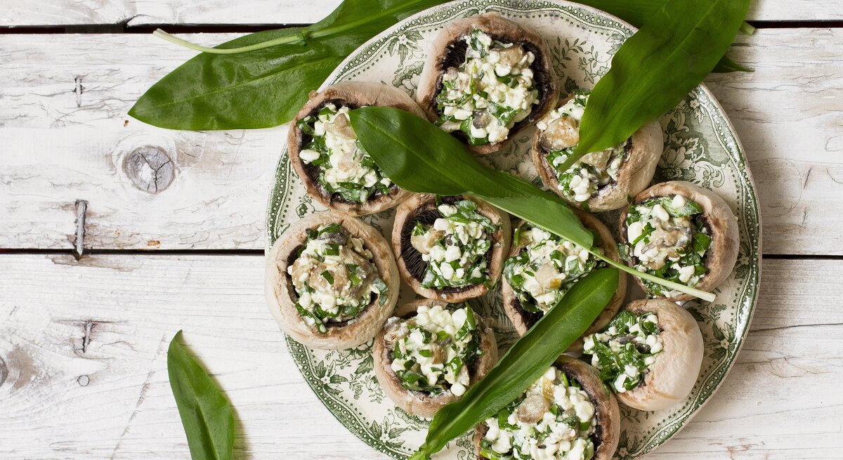 Gefüllte Champignons mit einer Füllung aus Bärlauch und Feta auf einem hellen verzierten Teller mit ganzen Bärlauch-Blättern auf einem hellen Holztisch von oben.