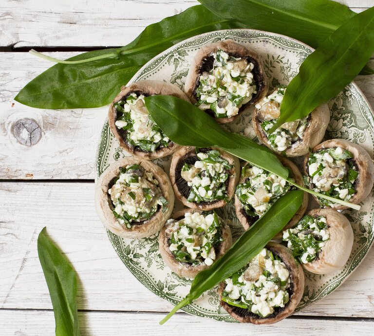 Gefüllte Champignons mit einer Füllung aus Bärlauch und Feta auf einem hellen verzierten Teller mit ganzen Bärlauch-Blättern auf einem hellen Holztisch von oben. | © GettyImages/ Adél Békefi