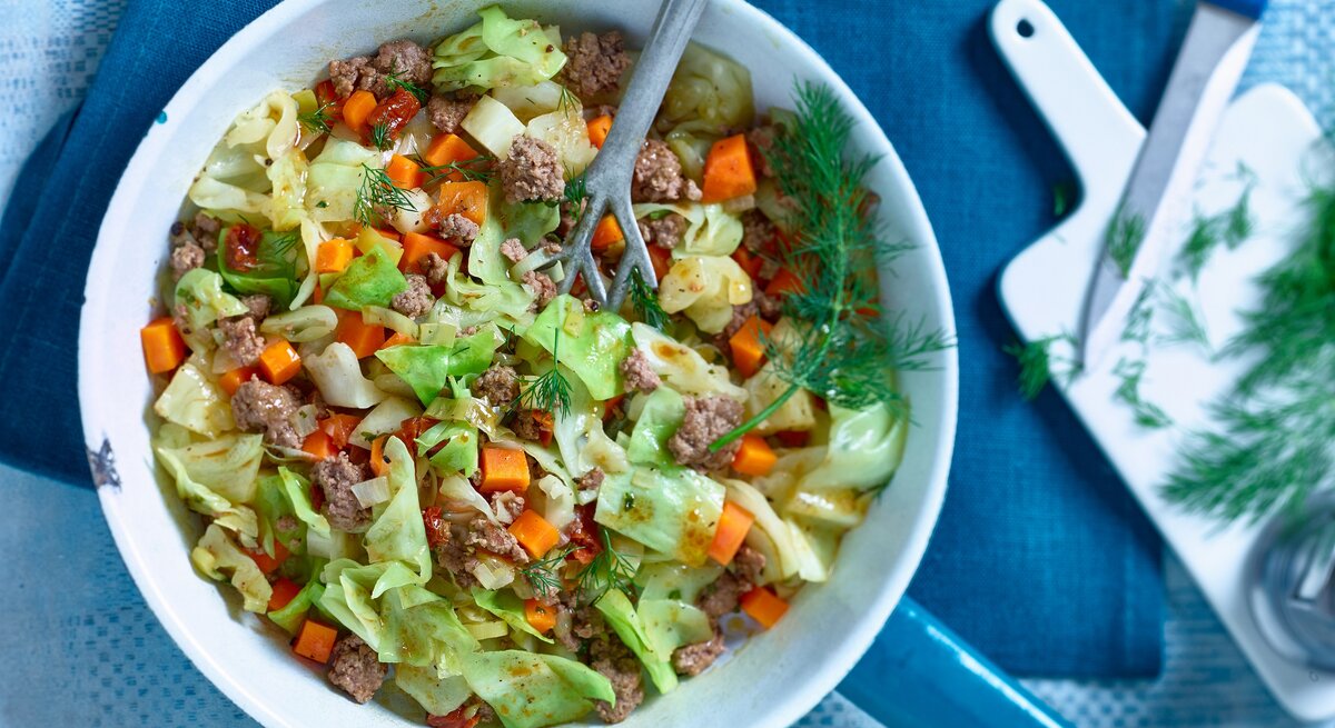 Spitzkohl-Hack-Pfanne mit Stil / Pfannenwender / Daneben Brettchen, Küchenmesser und frische Kräuter / Draufsicht
