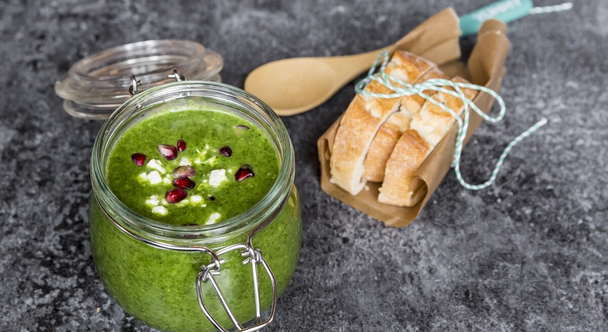 Grünkohlsuppe mit Granatapfel in einem Bügelverschlussglas vor rustikalem Hintergrund. Rechts Baguettte-Scheiben. Draufsicht.