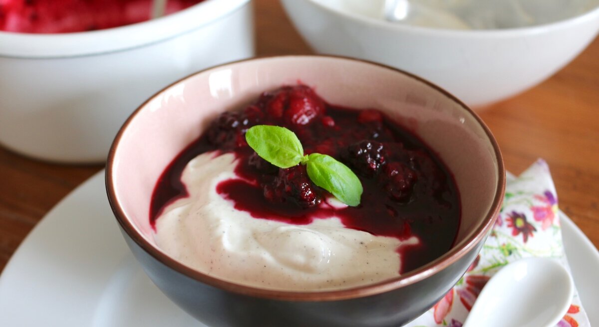 Quark-Dessert mit Vanille in einer dunklen Schüssel auf einem weißen Unterteller, rechts davon ein weißer Löffel und der Hintergrund ist hölzern.