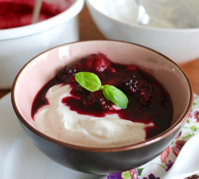 Quark-Dessert mit Vanille in einer dunklen Schüssel auf einem weißen Unterteller, rechts davon ein weißer Löffel und der Hintergrund ist hölzern. | © Nadine Hüttenrauch