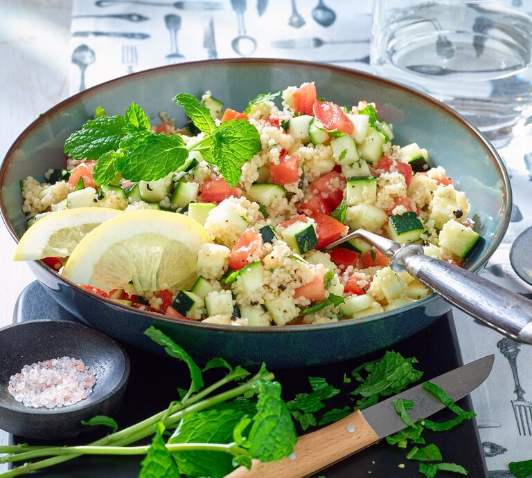 Couscous-Zucchini-Pfanne in einer blauen Schüssel. Auf dem Foto verteilt Salz, Gewürze, ein Messer und Wasser. | © Matthias Liebich