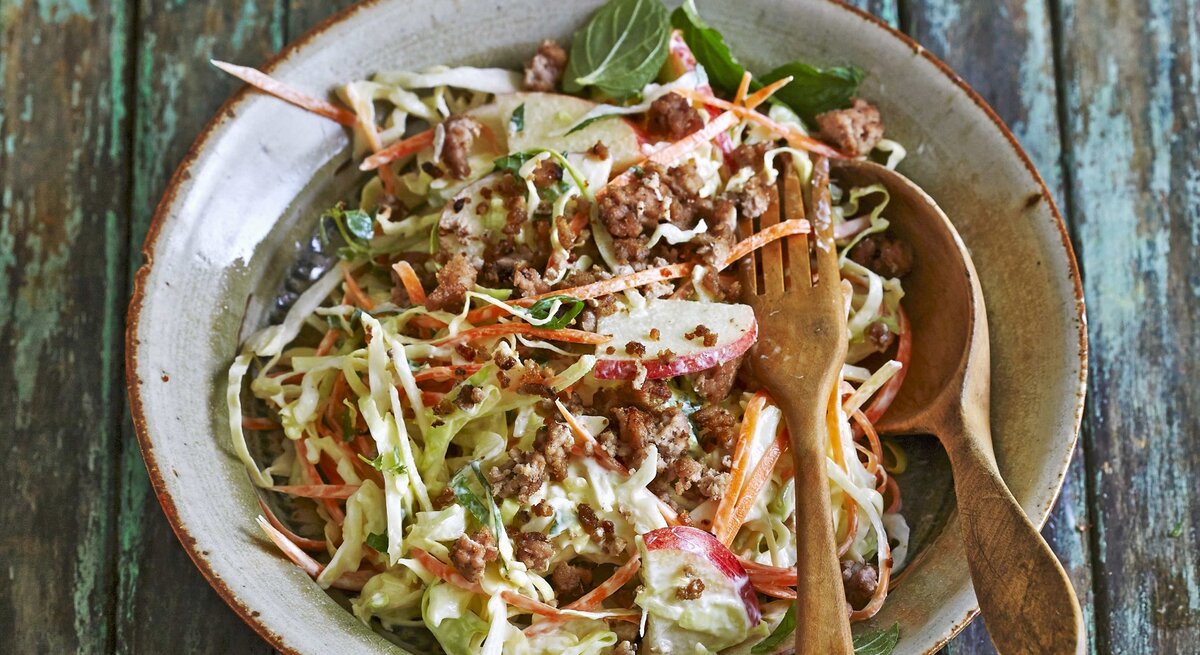 Möhren-Spitzkohl-Salat mit Rinderhack und Apfel / runder Teller / Löffel und Gabel aus Holz / Untergrund Holzplanken / Draufsicht, schräg