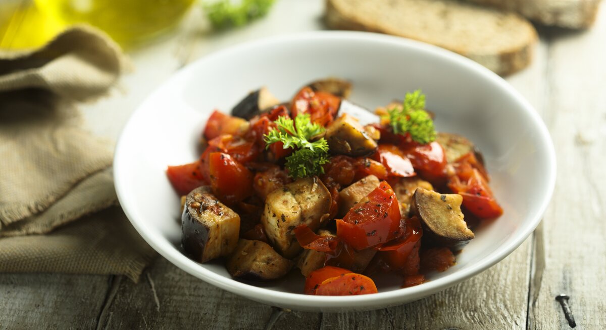 Italienischer Auberginen-Eintopf Caponata in weißem Teller auf rustikaler Oberfläche.