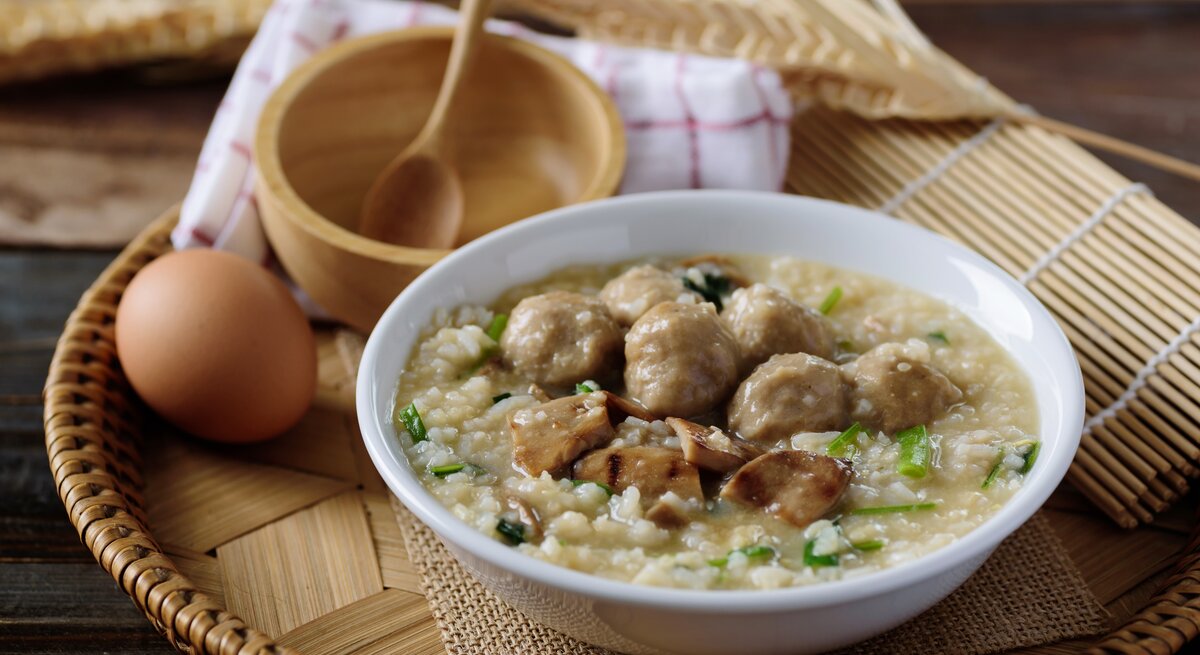 Reissuppe mit Hackbällchen & gebratenen Pilzen in weißer Schüssel auf hölzernem Untergrund.