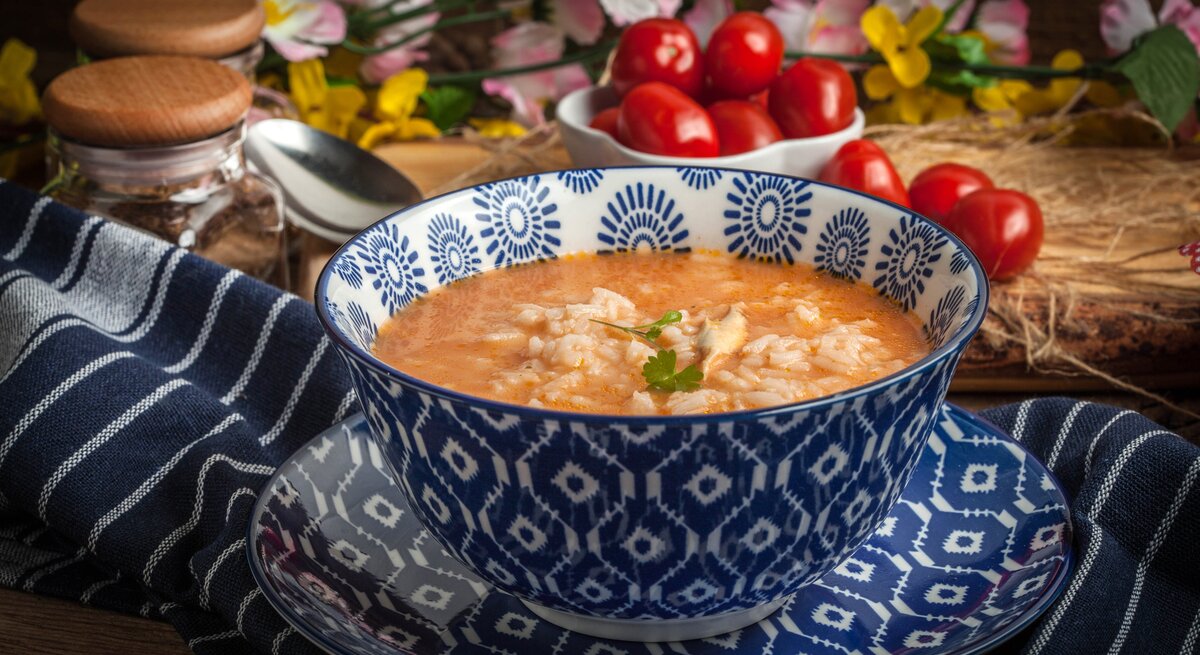 Eine Schüssel Tomatensuppe mit Reis, serviert in einer tiefblauen, mit weißen Mustern verzierten Schale auf einem Untersetzer gleichen Designs. Die Schale steht auf einem dunkelblauen Tuch mit weißen Streifen. Im Hintergrund sind Gewürzgläser, ein Löffel und frische Kirschtomaten zu sehen, umgeben von rustikalen Holzelementen und Frühlingsblumen.