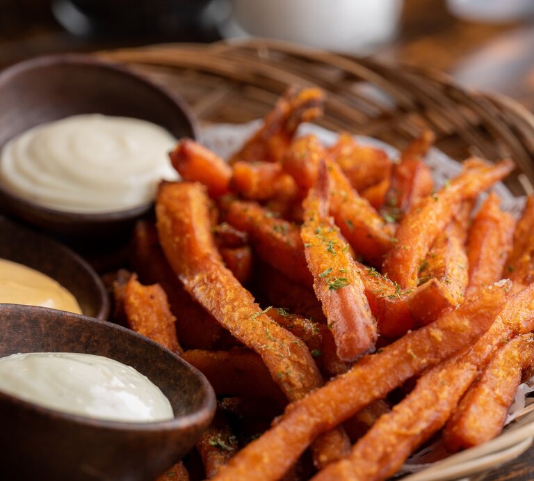 Süßkartoffelpommes aus der Heißluftfritteuse in Bambusschale mit dreierlei Dips in braunen Schalen auf Holztisch. Draufsicht. | © Getty Images / Justin Ong