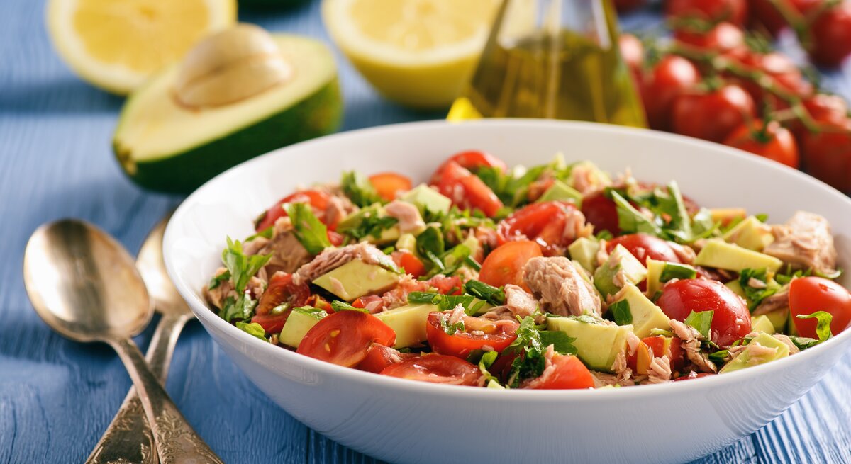 Weißer Teller mit Avocado-Thunfisch-Salat, Tomaten und Petersilie auf blauem Untergrund – umrundet von den Zutaten für den Salat.