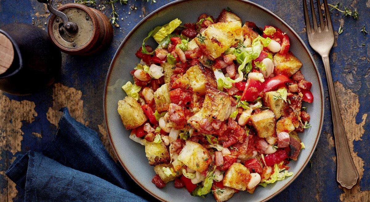 Ein grauer Teller mit dem Brotsalat mit Bohnen und Paprika auf dunkelblauem Holztisch direkt von oben fotografiert.