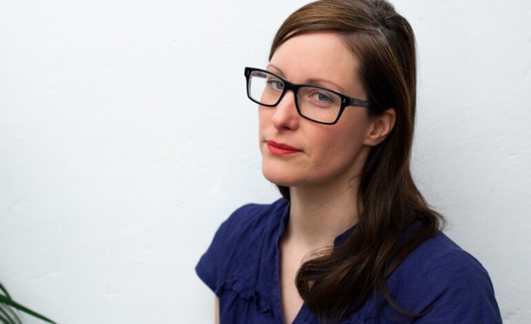 Eine Frau mit dunklen Haaren und Brille trägt ein blaues Oberteil und schaut nachdenklich zur Seite, vor einem weißen Hintergrund, was ein klares und einfaches Porträt schafft. | © privat