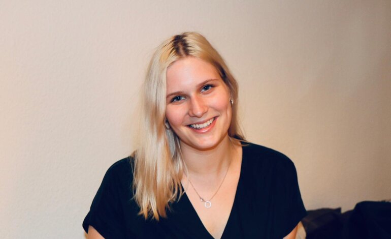 Das Bild zeigt eine junge Frau mit blonden Haaren, die in die Kamera lächelt. Sie trägt ein schwarzes Oberteil und eine Kette mit einem kleinen Anhänger. Ihr freundliches Lächeln und ihre offene Körperhaltung vermitteln eine positive und herzliche Ausstrahlung. Der Hintergrund ist neutral und unscharf, was den Fokus auf die Frau richtet. | © privat