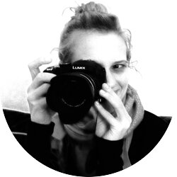 Schwarz-weiß Foto einer lächelnden Person, die eine Lumix Kamera vor ihr Gesicht hält, um ein Foto zu machen, wobei nur ein Auge sichtbar ist, was eine lebendige und kreative Atmosphäre schafft. | © privat