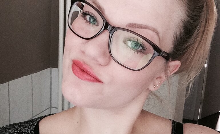 Selbstporträt einer Frau mit zurückgebundenen blonden Haaren und großer Brille, die leuchtend roten Lippenstift trägt und direkt in die Kamera blickt. | © privat