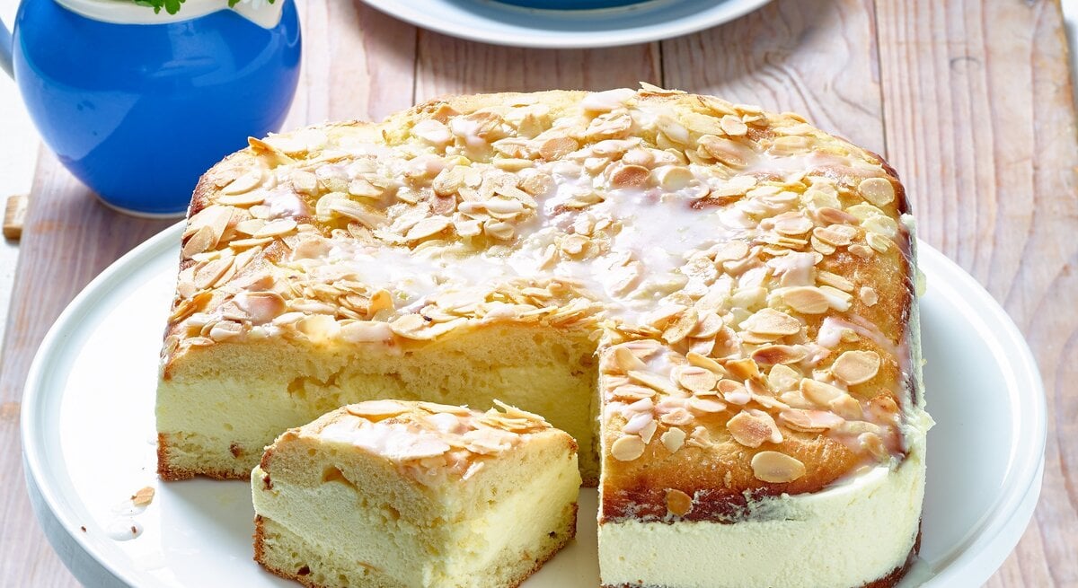 Der Bienenstich mit Zitronen-Mousse hat eine cremige Füllung und einen luftigen Teig. Die obere Schicht des Kuchens ist mit gerösteten Mandelsplittern und einem Zuckerguss bedeckt. Ein Stück des Kuchens wurde herausgeschnitten und liegt neben dem ganzen Kuchen auf der Tortenplatte. Im Hintergrund steht eine kleine Vase mit Blumen und ein Teller mit einem weiteren Stück Kuchen. Der Tisch ist rustikal aus Holz.