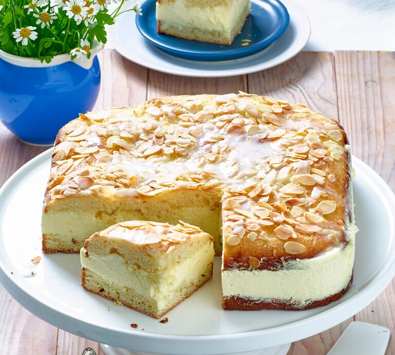Der Bienenstich mit Zitronen-Mousse hat eine cremige Füllung und einen luftigen Teig. Die obere Schicht des Kuchens ist mit gerösteten Mandelsplittern und einem Zuckerguss bedeckt. Ein Stück des Kuchens wurde herausgeschnitten und liegt neben dem ganzen Kuchen auf der Tortenplatte. Im Hintergrund steht eine kleine Vase mit Blumen und ein Teller mit einem weiteren Stück Kuchen. Der Tisch ist rustikal aus Holz. | © Matthias Liebich