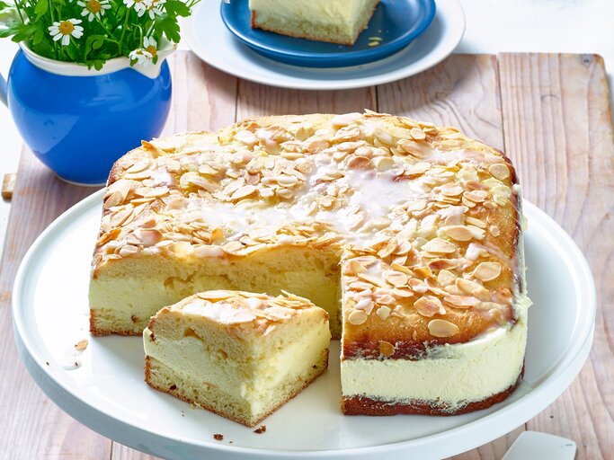 Der Bienenstich mit Zitronen-Mousse hat eine cremige Füllung und einen luftigen Teig. Die obere Schicht des Kuchens ist mit gerösteten Mandelsplittern und einem Zuckerguss bedeckt. Ein Stück des Kuchens wurde herausgeschnitten und liegt neben dem ganzen Kuchen auf der Tortenplatte. Im Hintergrund steht eine kleine Vase mit Blumen und ein Teller mit einem weiteren Stück Kuchen. Der Tisch ist rustikal aus Holz. | © Matthias Liebich