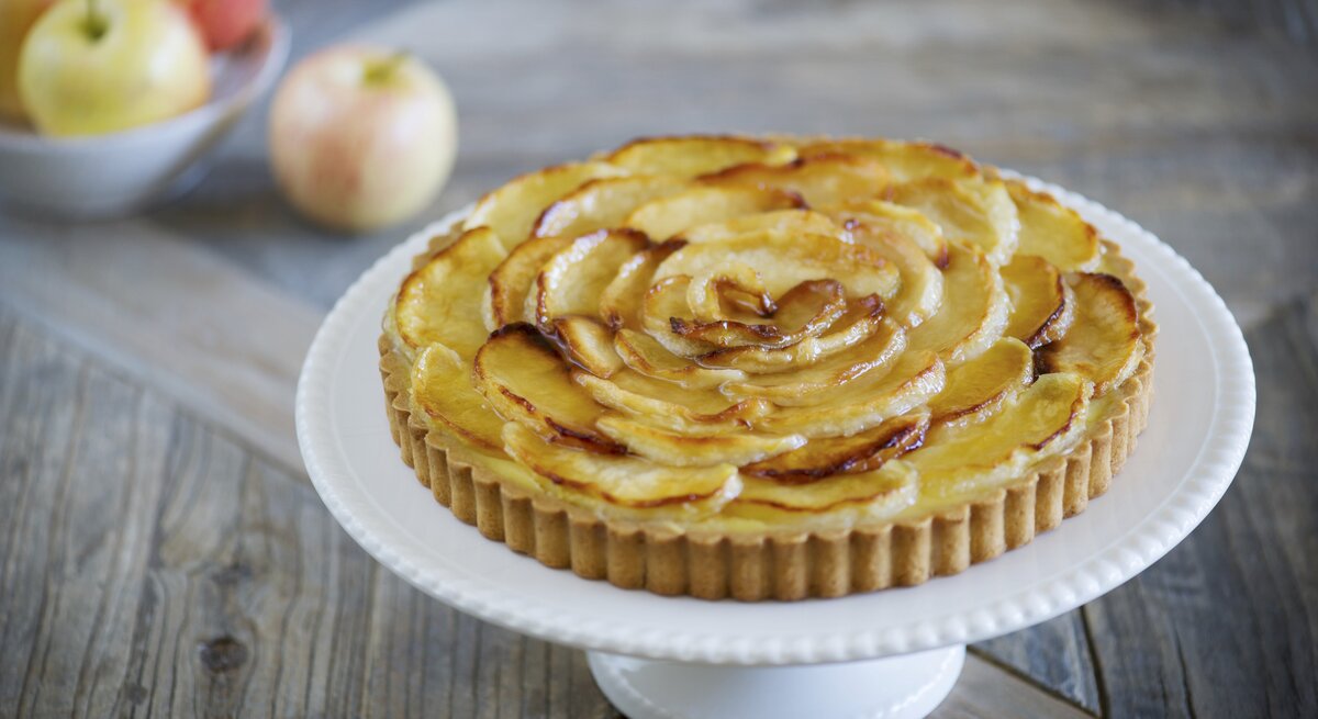 Die Apfel-Tarte auf einem weißen Tortenständer und rustikalem Holztisch von schräg oben fotografiert.