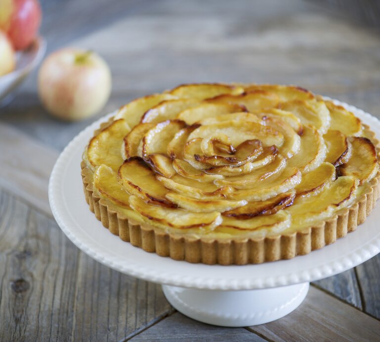 Die Apfel-Tarte auf einem weißen Tortenständer und rustikalem Holztisch von schräg oben fotografiert. | © StockFood / Beller, Gaelle