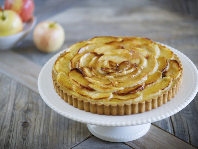 Die Apfel-Tarte auf einem weißen Tortenständer und rustikalem Holztisch von schräg oben fotografiert. | © StockFood / Beller, Gaelle