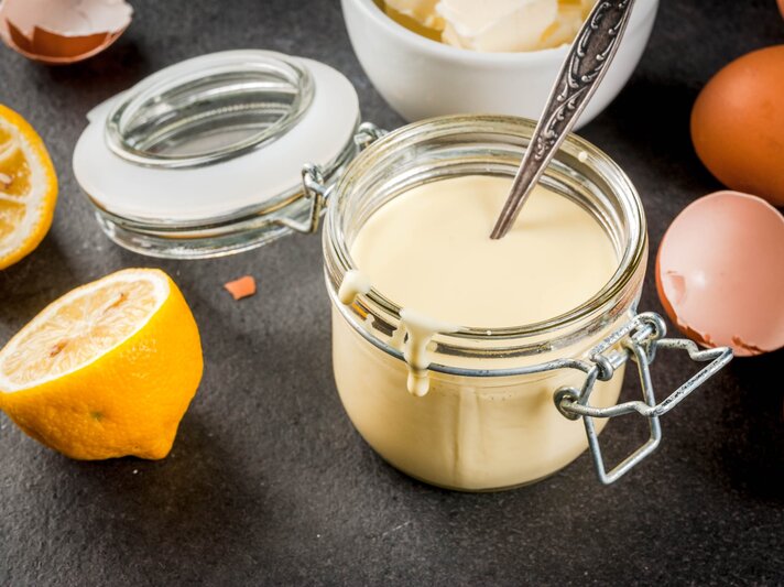 Ein Weckglas mit der Sauce hollandaise auf einem grauen Untergrund. Alles von schräg oben fotografiert und im Hintergrund sind noch die Zutaten erkennbar. | © iStock/Rimma_Bondarenko