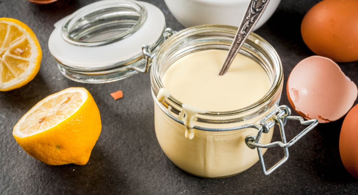 Ein Weckglas mit der Sauce hollandaise auf einem grauen Untergrund. Alles von schräg oben fotografiert und im Hintergrund sind noch die Zutaten erkennbar.