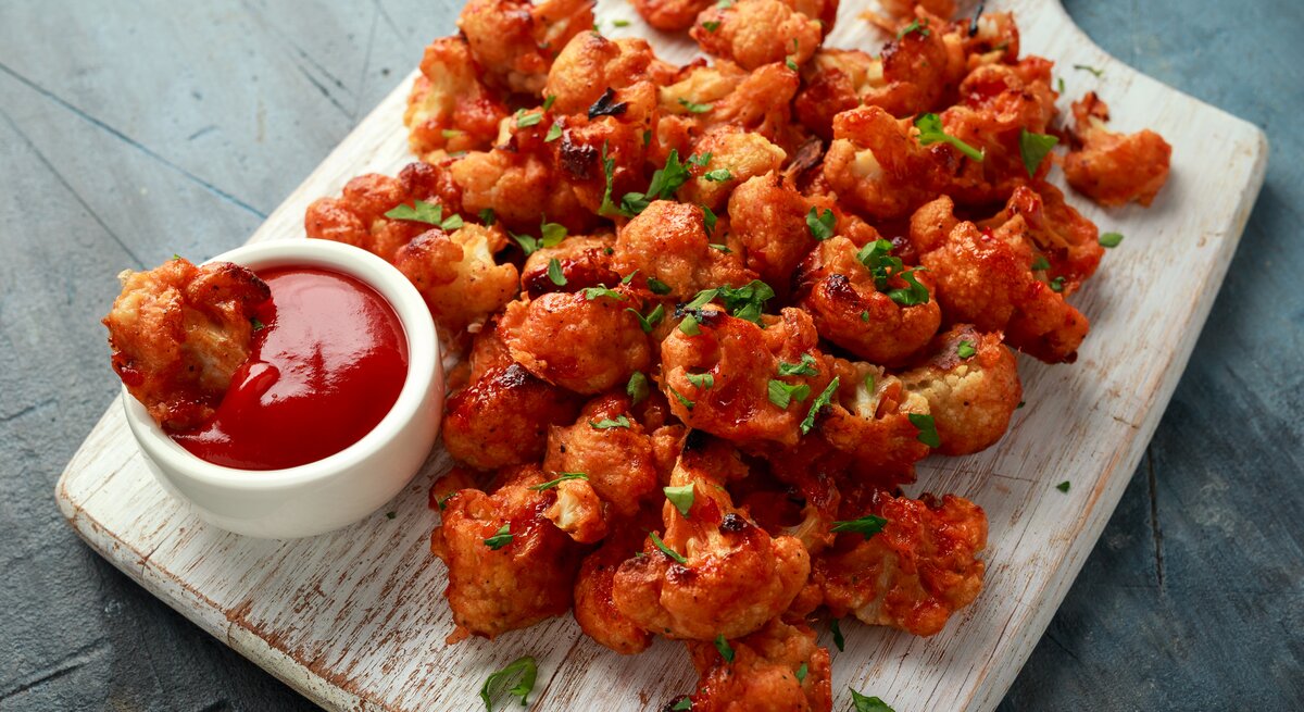 Blumenkohl Wings in scharfer Soße liegen auf einem Holzbrett auf einem grauen Untergrund. Neben den Wings steht eine kleine Schüssel mit scharfer Sauce.