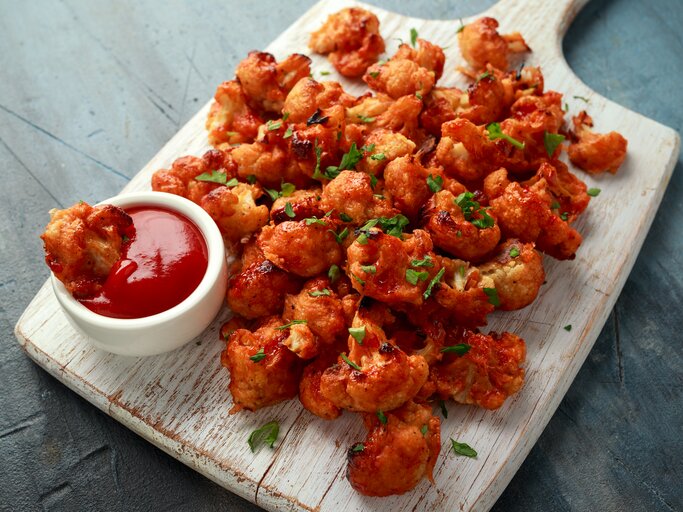 Blumenkohl Wings in scharfer Soße liegen auf einem Holzbrett auf einem grauen Untergrund. Neben den Wings steht eine kleine Schüssel mit scharfer Sauce. | © Getty Images / DronG