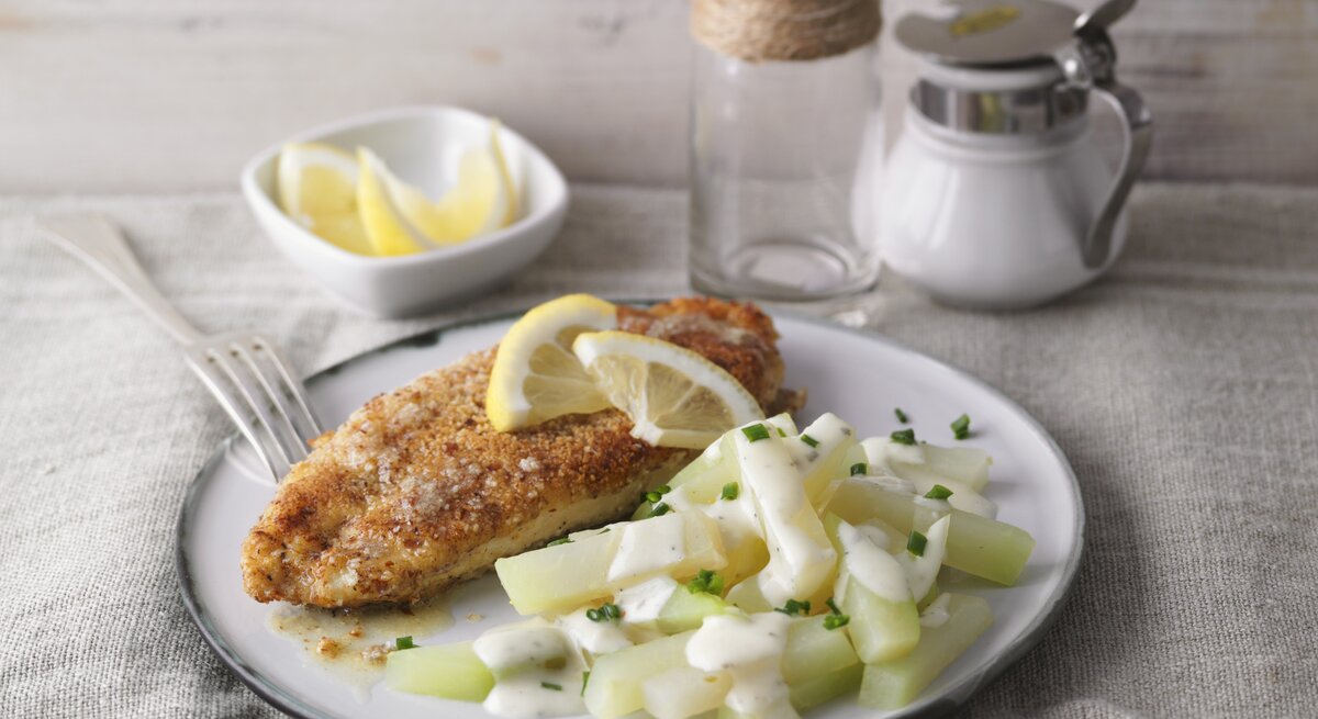 Ein weißer Teller mit dem Mandel-Schnitzel und Kohlrabi-Rahm auf einem hellen Tisch mit Zitronenecken garniert.