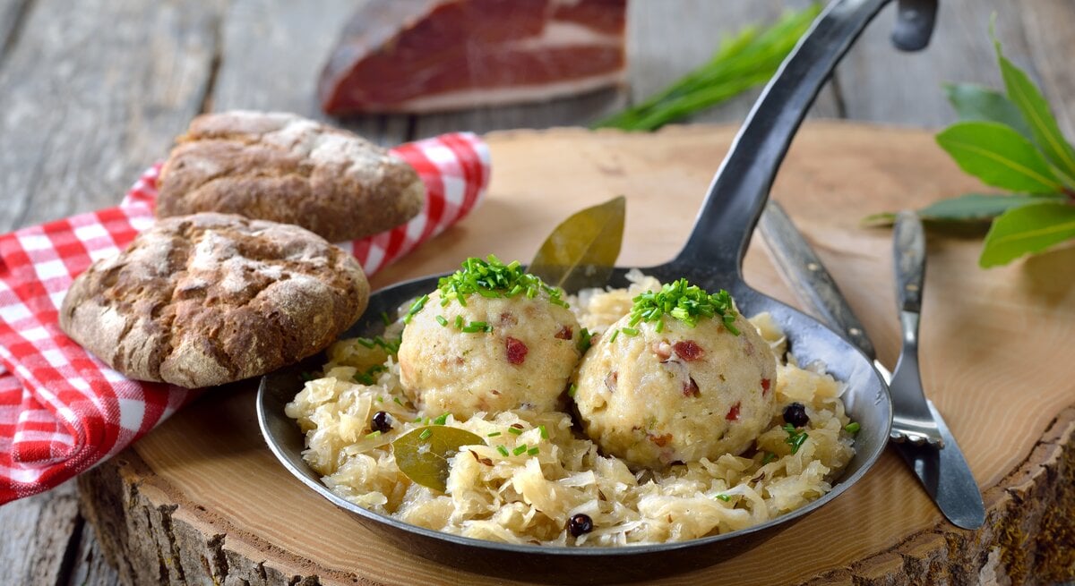 Ein Baumstamm steht auf einem Untergrund aus Holz. Darauf steht eine schmiedeeiserne Pfanne mit Sauerkraut und  selbst gemachten Speckknödeln. Daneben 