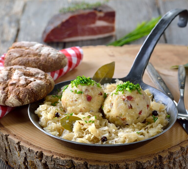 Ein Baumstamm steht auf einem Untergrund aus Holz. Darauf steht eine schmiedeeiserne Pfanne mit Sauerkraut und  selbst gemachten Speckknödeln. Daneben  | © Getty Images / kabVisio