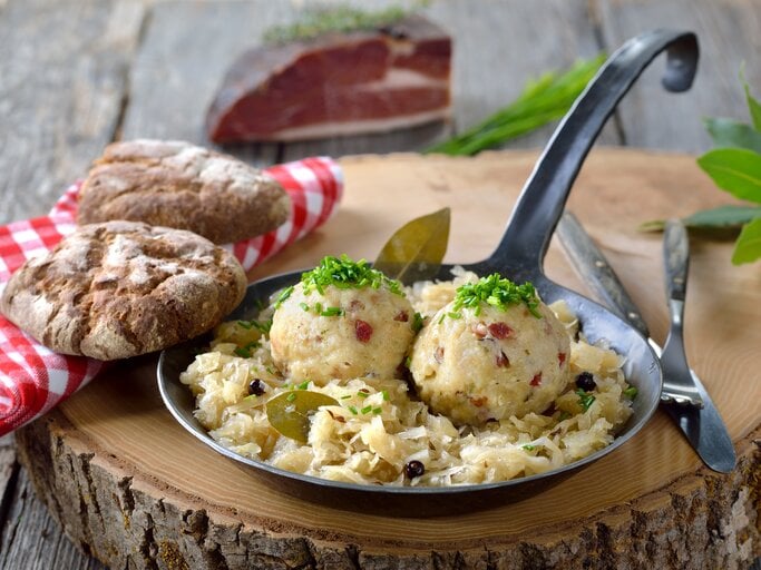 Ein Baumstamm steht auf einem Untergrund aus Holz. Darauf steht eine schmiedeeiserne Pfanne mit Sauerkraut und  selbst gemachten Speckknödeln. Daneben  | © Getty Images / kabVisio