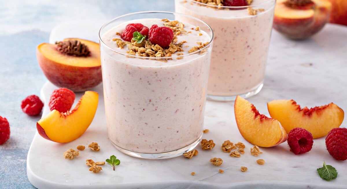 Frontal: Zwei Himbeer-Nektarinen-Smoothie im Glas stehen auf einem weißen Marmorbrettchen, das auf einem hellen Untergund liegt. Drum herum liegen aufgeschnitten Nekatarinenscheiben und frische Himmbeeren.