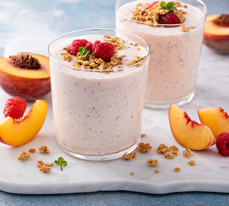 Frontal: Zwei Himbeer-Nektarinen-Smoothie im Glas stehen auf einem weißen Marmorbrettchen, das auf einem hellen Untergund liegt. Drum herum liegen aufgeschnitten Nekatarinenscheiben und frische Himmbeeren. | © Getty Images / 	VeselovaElena