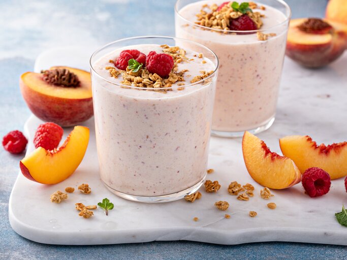 Frontal: Zwei Himbeer-Nektarinen-Smoothie im Glas stehen auf einem weißen Marmorbrettchen, das auf einem hellen Untergund liegt. Drum herum liegen aufgeschnitten Nekatarinenscheiben und frische Himmbeeren. | © Getty Images / 	VeselovaElena