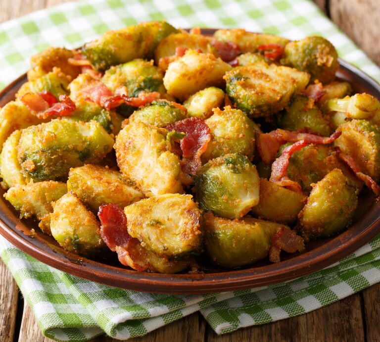 Brauner Teller belegt mit gebratenem Rosenkohl, Speck und Semmelbröseln. Das Ganze steht auf einem Holztisch.  | © iStock/ALLEKO