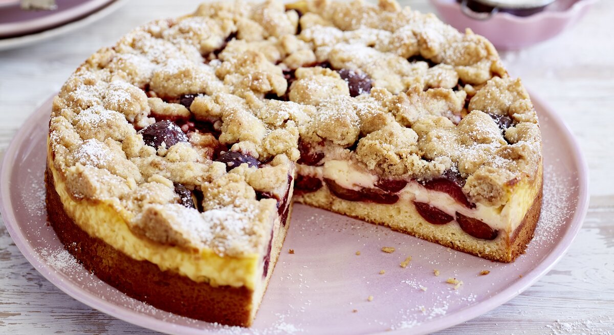 Ein rosa Teller mit einem Stück des Pflaumen-Puddingkuchens und Zimtstreuseln auf einem hellen Holztisch.