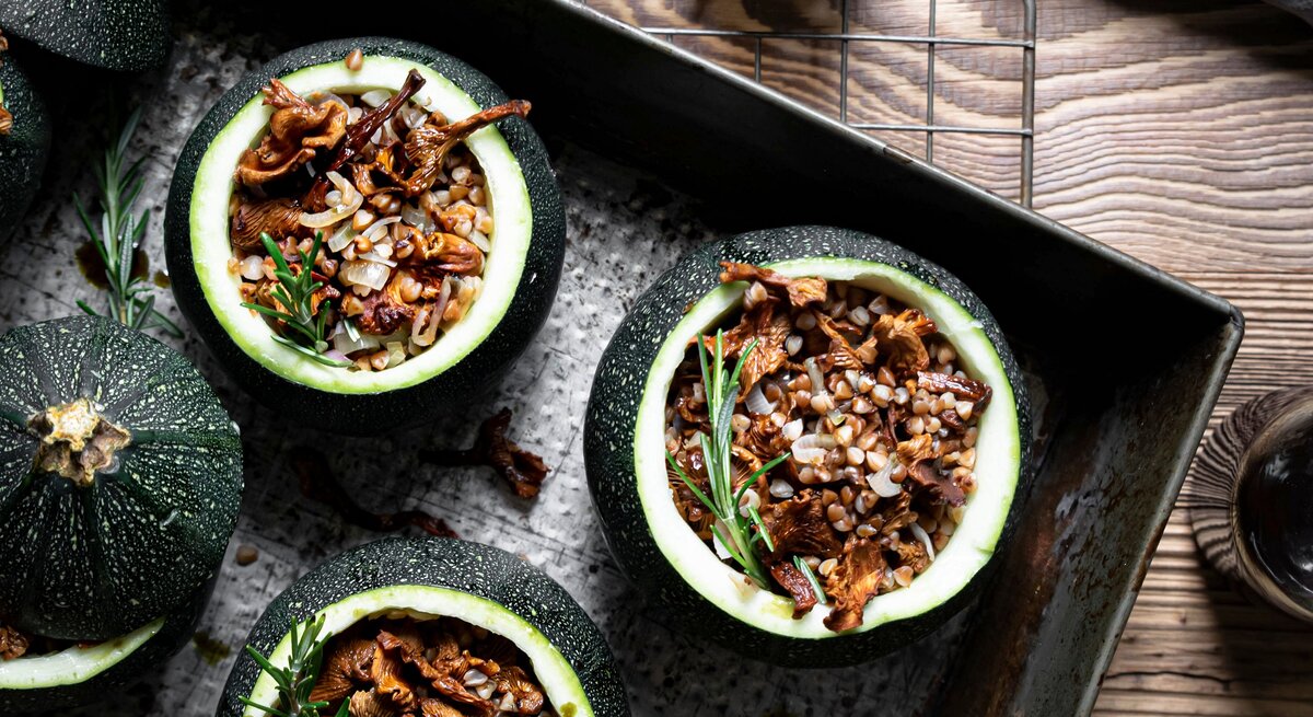 Draufsicht: Vier runde gefüllte Zucchini mit Pfifferlingen und Buchweizen auf einem dunklem Tablett, dass auf einem Holztisch liegt. Daneben steht ein Pfeffermühle sowie ein Schale mit der Zucchini-Füllung.