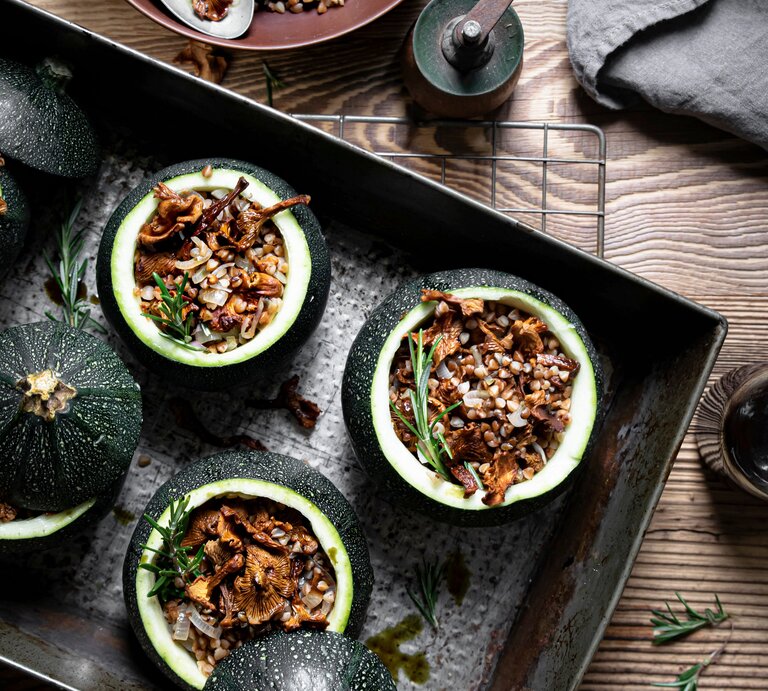 Draufsicht: Vier runde gefüllte Zucchini mit Pfifferlingen und Buchweizen auf einem dunklem Tablett, dass auf einem Holztisch liegt. Daneben steht ein Pfeffermühle sowie ein Schale mit der Zucchini-Füllung. | © Shutterstock / Rawpixel.com