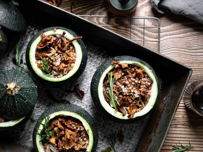 Draufsicht: Vier runde gefüllte Zucchini mit Pfifferlingen und Buchweizen auf einem dunklem Tablett, dass auf einem Holztisch liegt. Daneben steht ein Pfeffermühle sowie ein Schale mit der Zucchini-Füllung. | © Shutterstock / Rawpixel.com