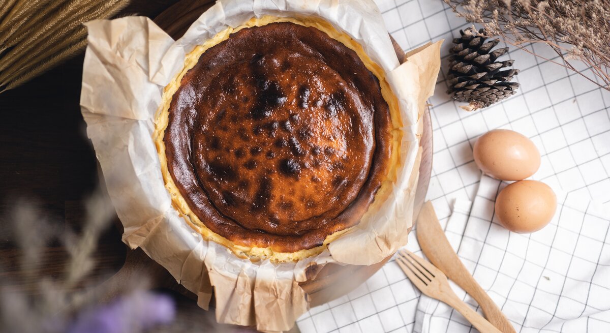 Frisch gebackener, leicht verbrannter Käsekuchen auf Backpapier, umgeben von Eiern und Naturdeko auf karierter Stoffserviette.