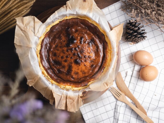 Frisch gebackener, leicht verbrannter Käsekuchen auf Backpapier, umgeben von Eiern und Naturdeko auf karierter Stoffserviette. | © iStock.com/Skazzjy
