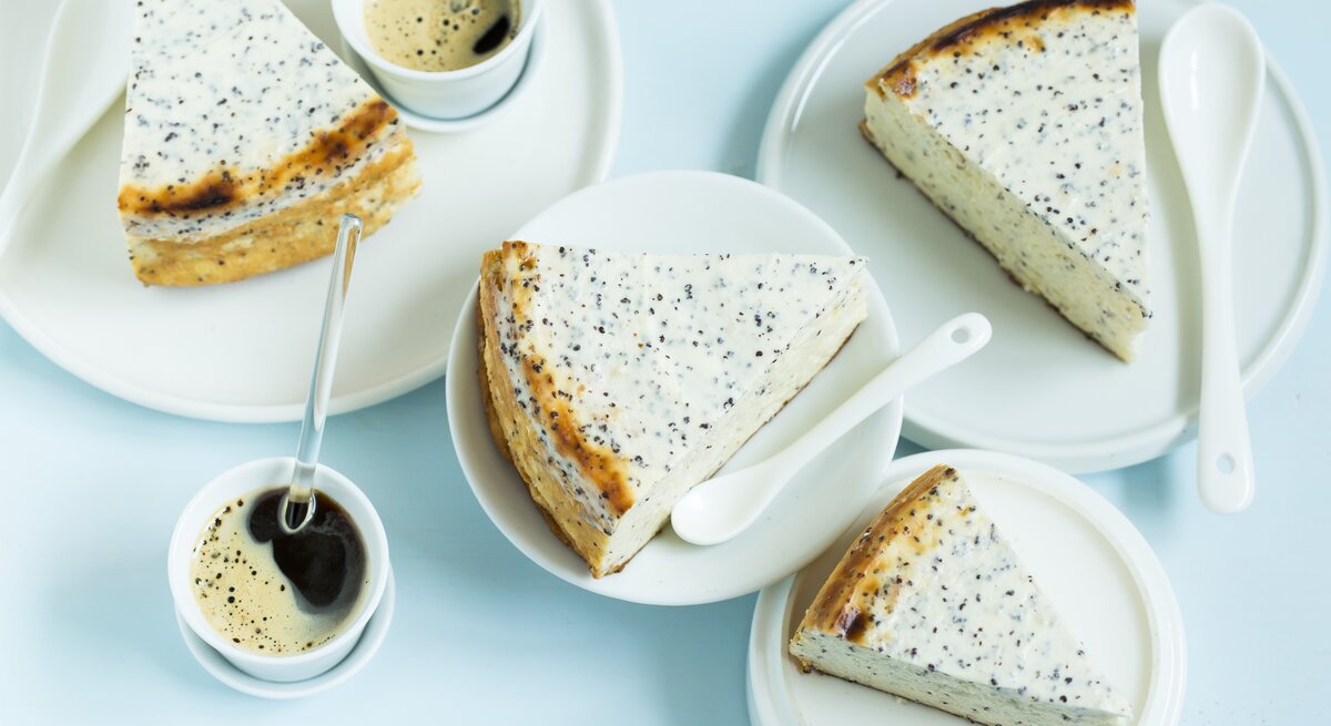 Mehrere Stücken vom Käsekuchen auf weißem Geschirr und einer hellblauen Tischdecke. Daneben noch Kaffee in kleinen Tassen. Alles direkt von oben fotografiert.