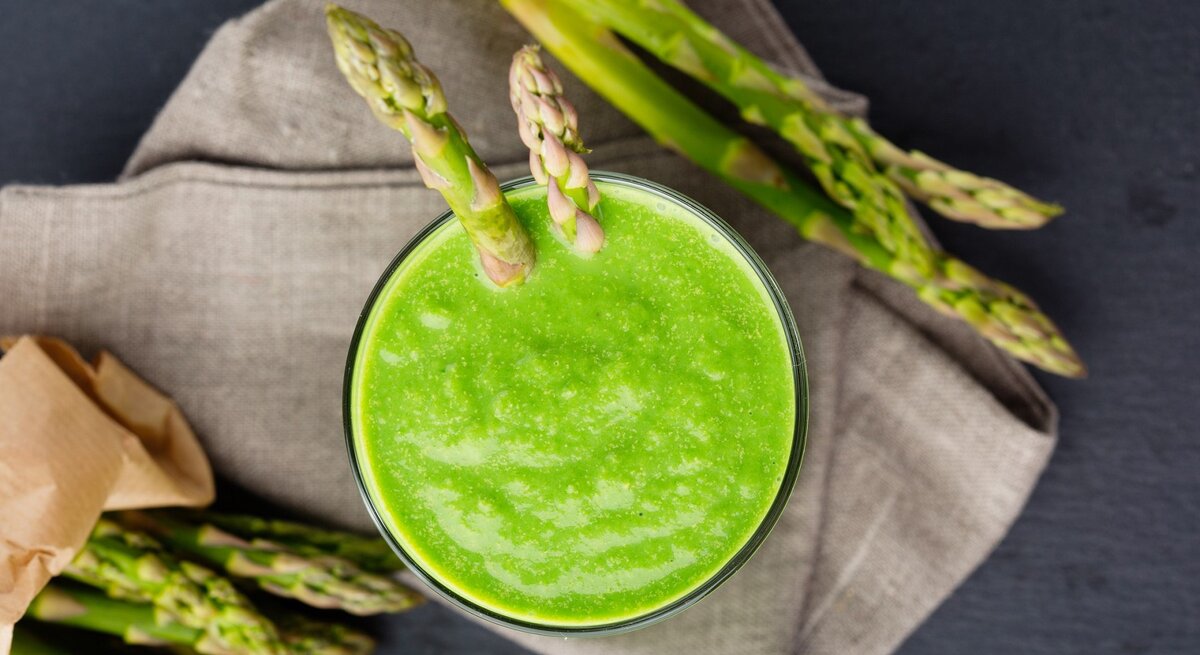 Grüner Smoothie mit Spargel in einem Glas. Darunter ein Küchentuch und daneben noch ein paar grüne Spargelstangen. 