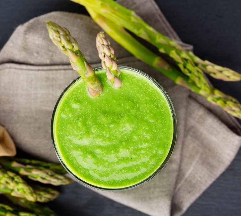 Grüner Smoothie mit Spargel in einem Glas. Darunter ein Küchentuch und daneben noch ein paar grüne Spargelstangen.  | © GettyImages/Metkalova