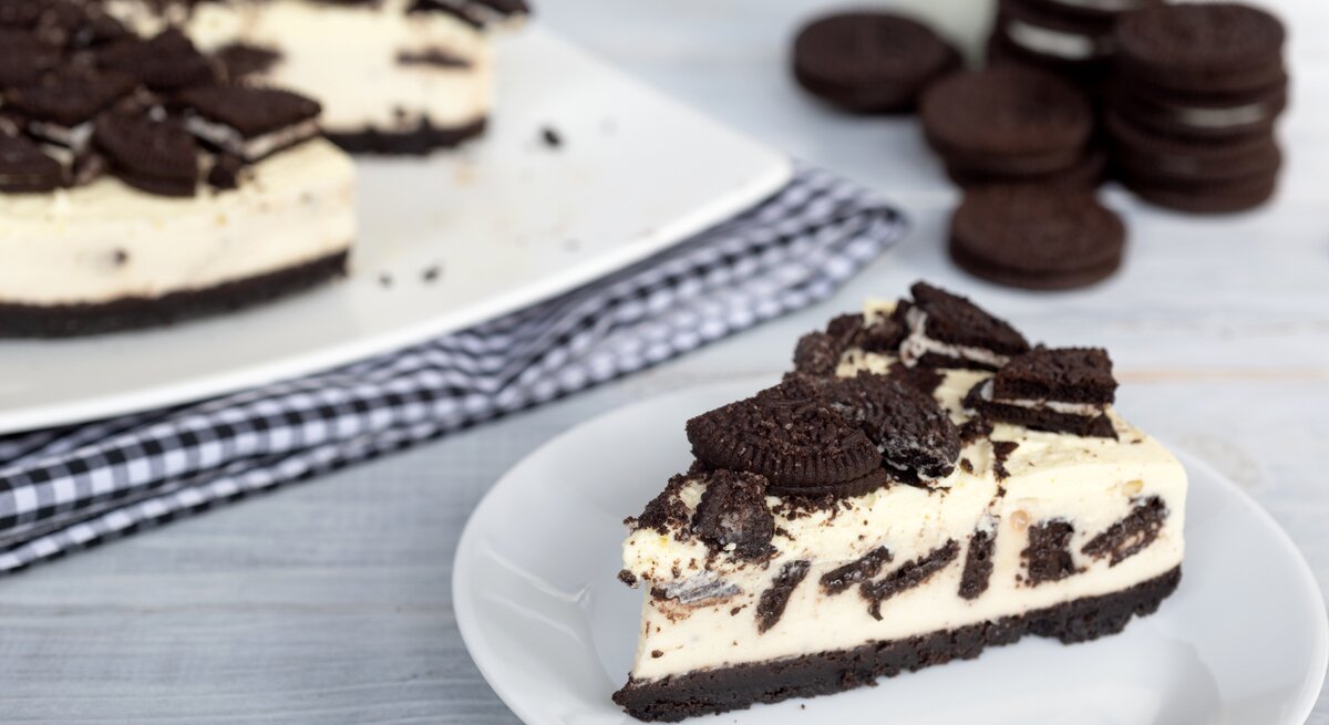 Ein Stück cremiger Oreo-Kuchen auf weißem Teller, daneben der restliche Kuchen auf kariertem Tuch und gestapelte Oreos im Hintergrund.