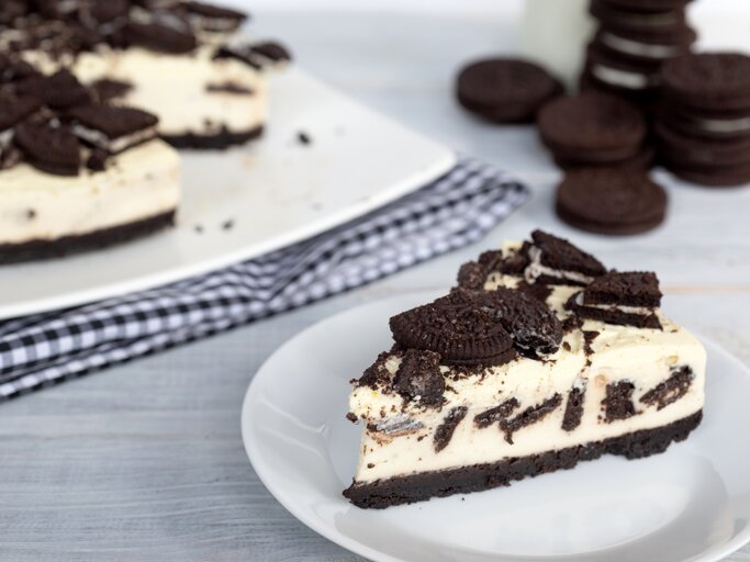 Ein Stück cremiger Oreo-Kuchen auf weißem Teller, daneben der restliche Kuchen auf kariertem Tuch und gestapelte Oreos im Hintergrund. | © iStock.com/Premyuda Yospim