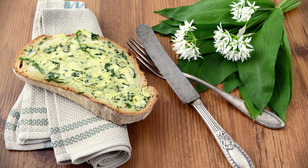 Brotscheibe beschmiert mit Bärlauchbutter auf Holztisch, daneben Besteck.