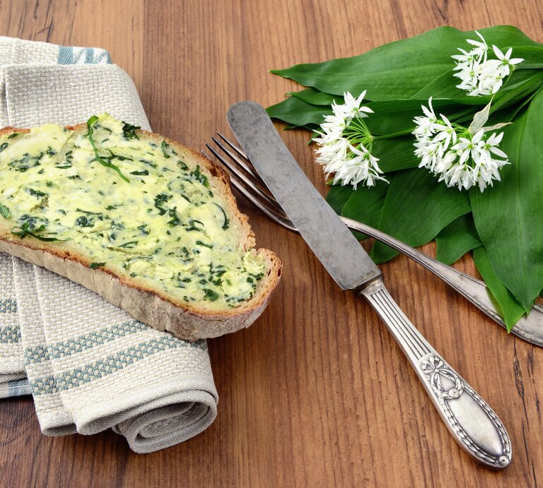 Brotscheibe beschmiert mit Bärlauchbutter auf Holztisch, daneben Besteck. | © iStock/hsvrs