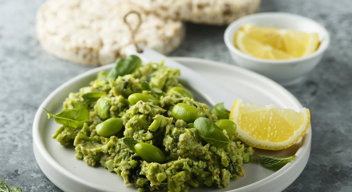 Frontal: eine weiße Schale Edamame-Guacamole steht auf einem grauen Untergrund. Sie ist mit einer Zitronenscheibe garniert. Im Hintergrund liegen Reiswaffeln und noch mehr Zitronenscheiben.